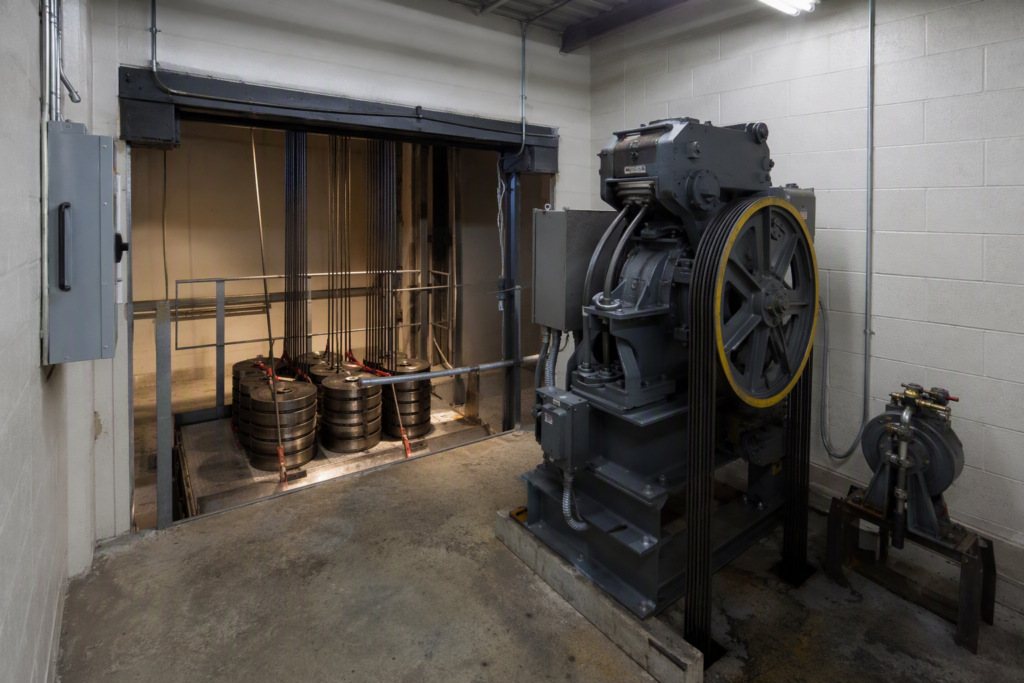 Elevator load test in progress in a Los Angeles commercial building machine room, showing calibrated test weights on the car roof and hoisting machinery